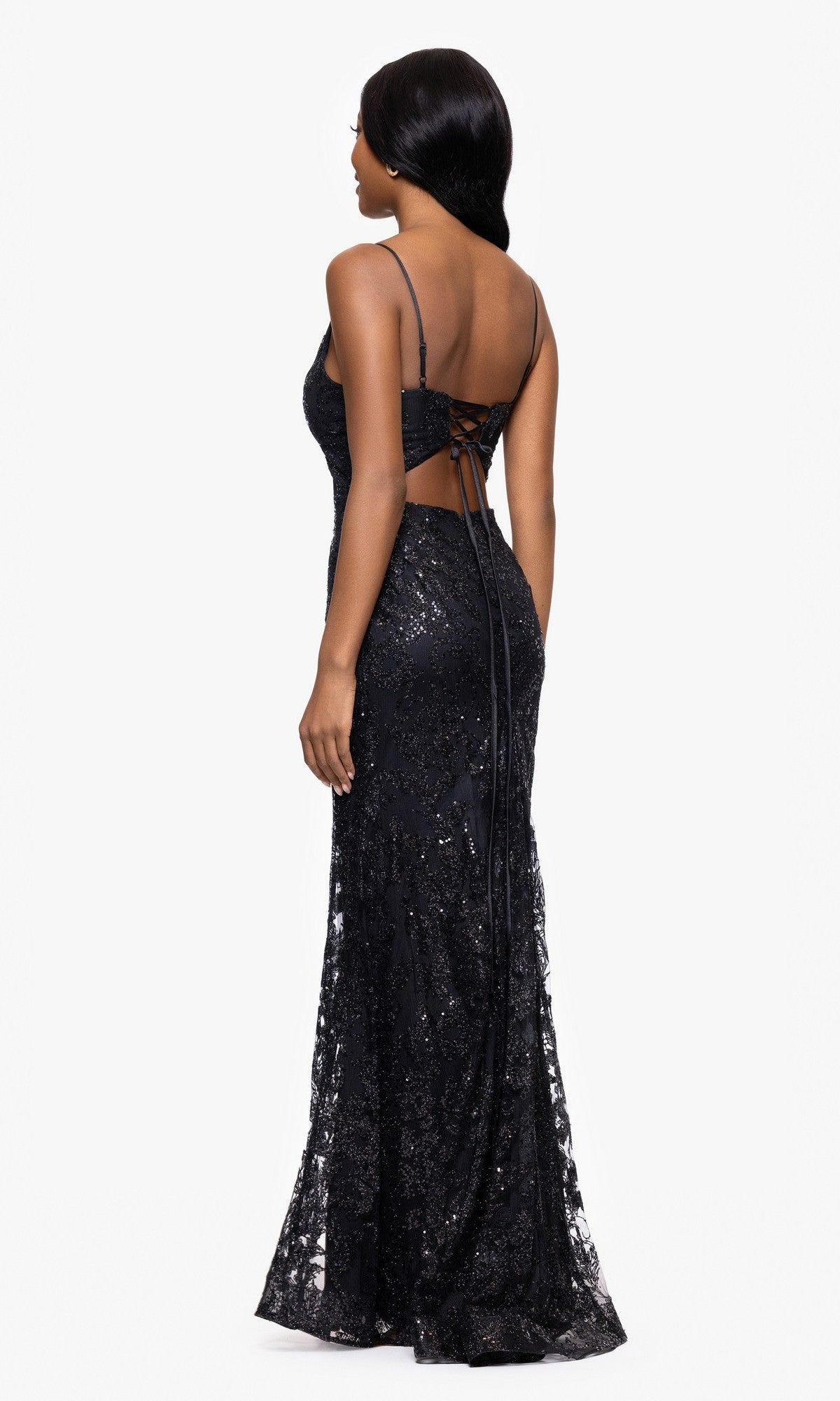 A woman stands facing away, wearing the Blondie Nites 4501BN—a long sequin-print prom dress with thin straps, open back, and lace-up detailing.